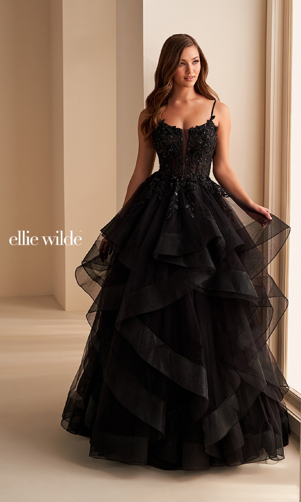 A woman wearing the Ellie Wilde EW36225, a layered black ball gown with a fitted, embellished bodice, stands near a window; this long prom dress exudes stunning designer elegance.